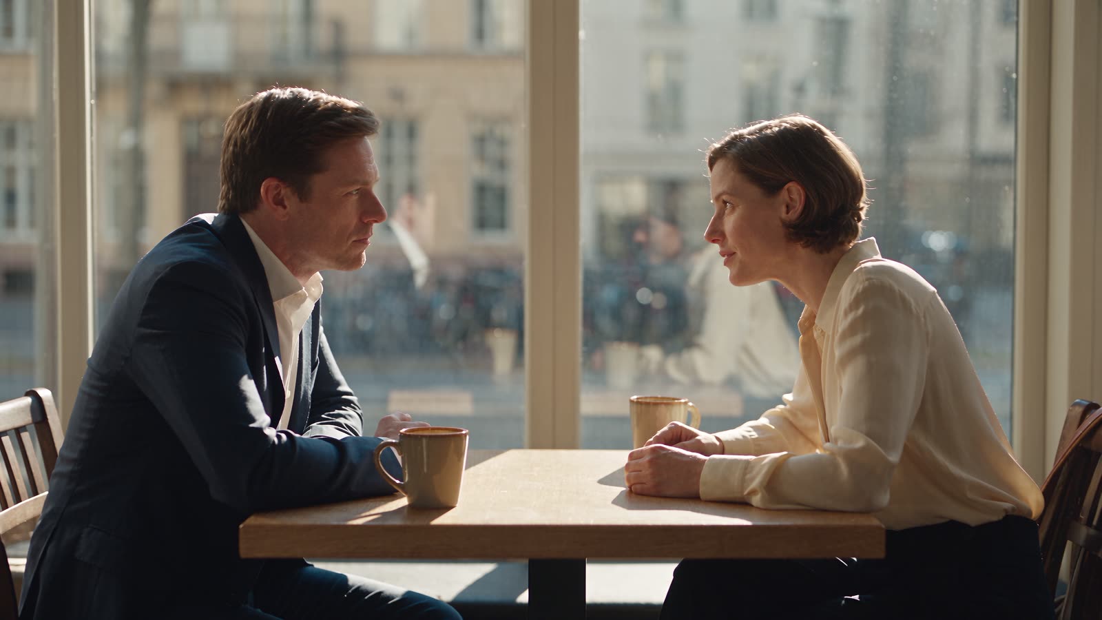 Two people in conversation across a cafe table by a sunlit window, coffee cups between them.